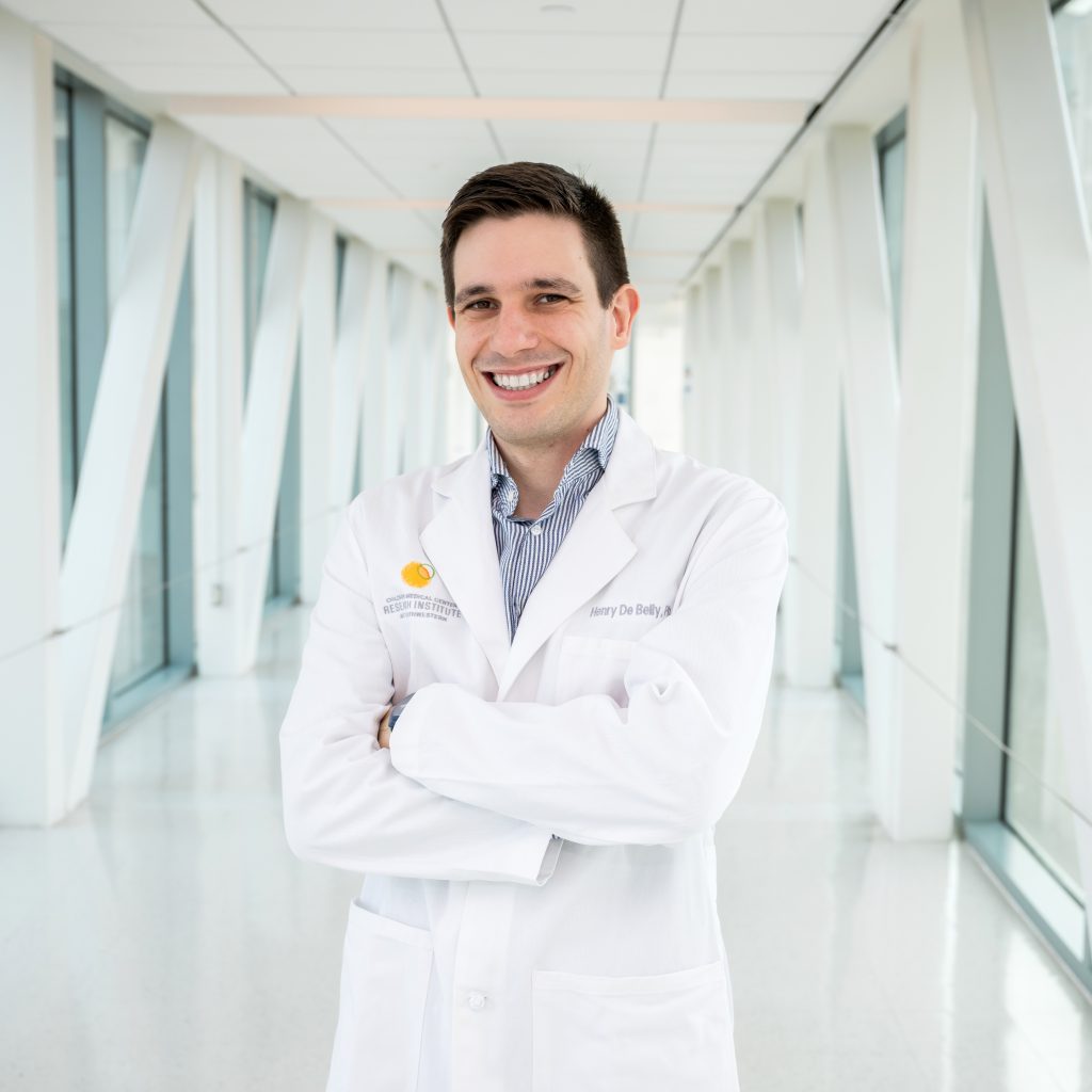 Henry De Belly stands in a hallway with windows, with his arms crossed, in a white lab coat.
